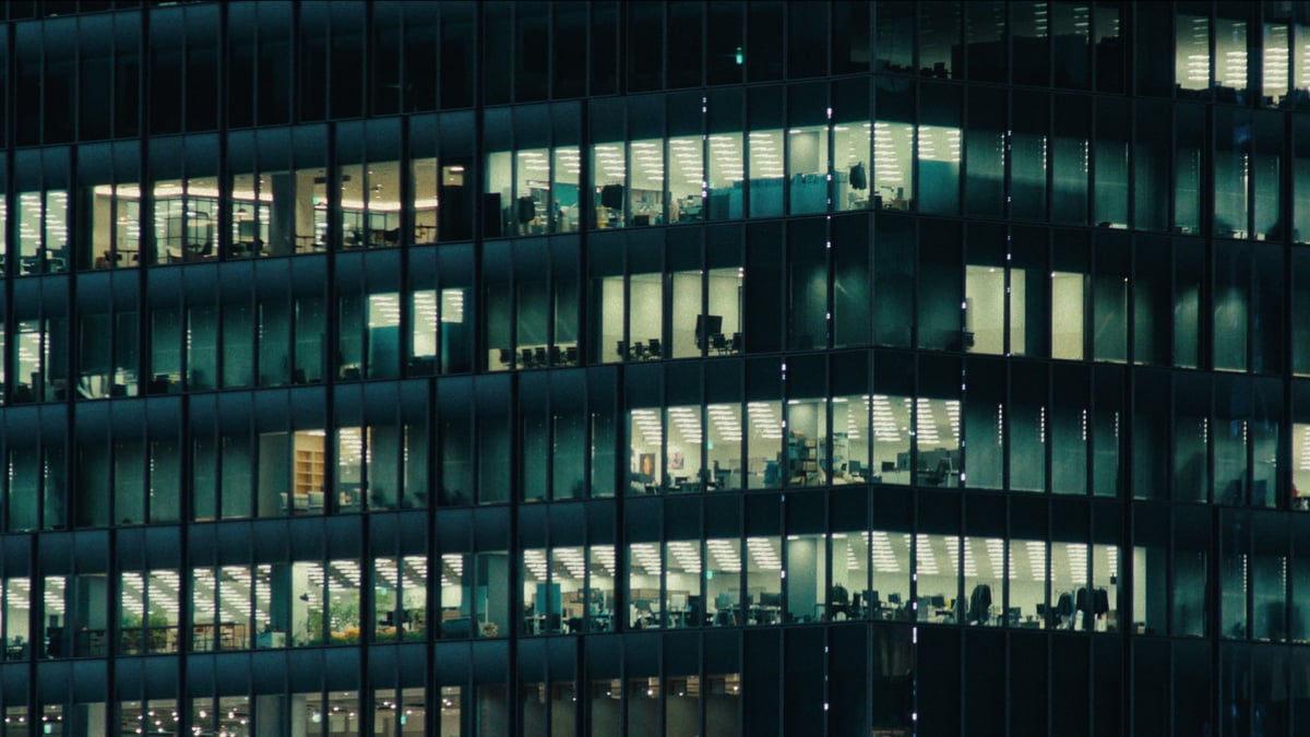 A nighttime exterior of a glass office tower with illuminated floors revealing empty workstations and meeting rooms.