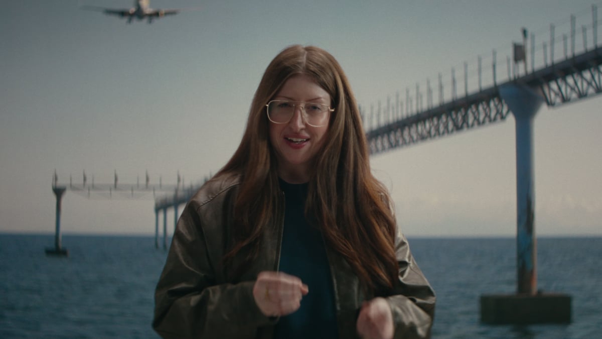 A woman with long auburn hair and oversized aviator glasses speaks to camera near the ocean as a plane descends behind her toward a pier.