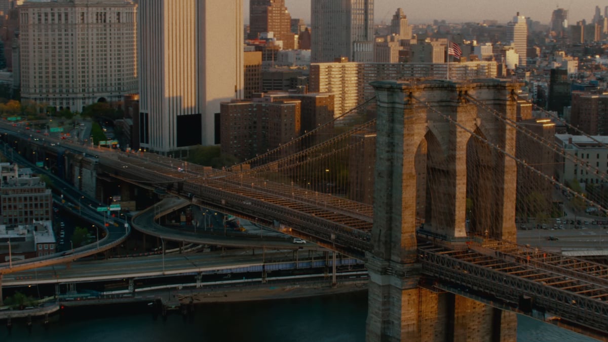 A sweeping aerial view of the Brooklyn Bridge at sunset, with its stone towers and cables in the foreground and the Manhattan skyline glowing in warm light behind.