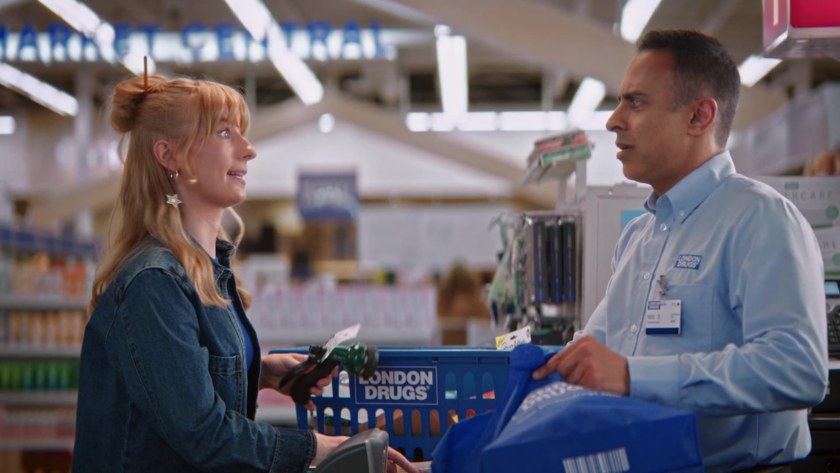 A London Drugs cashier hands over a shopping bag to a customer at checkout.