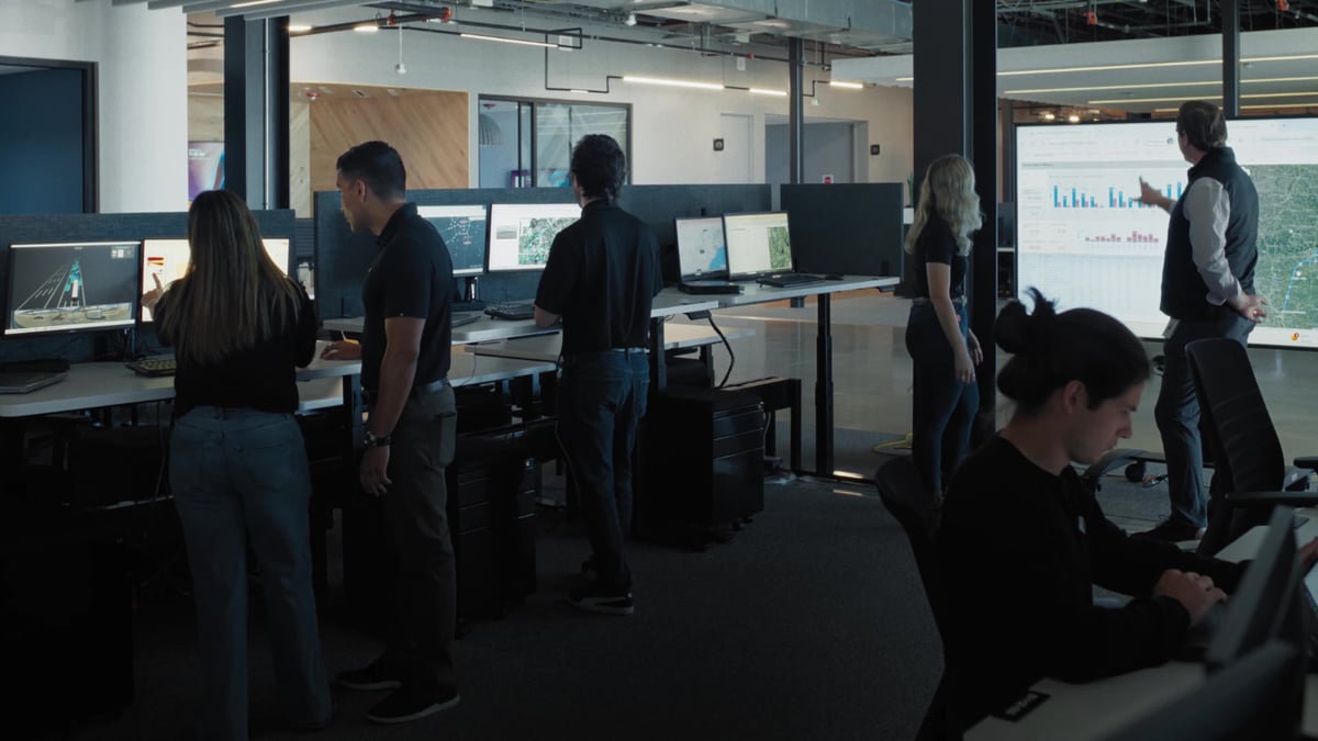 A team of technicians work at multiple monitor stations in a modern operations center, reviewing maps and data on large screens