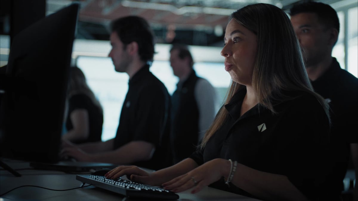 A woman in a black uniform works at a computer workstation in a busy operations center alongside her colleagues