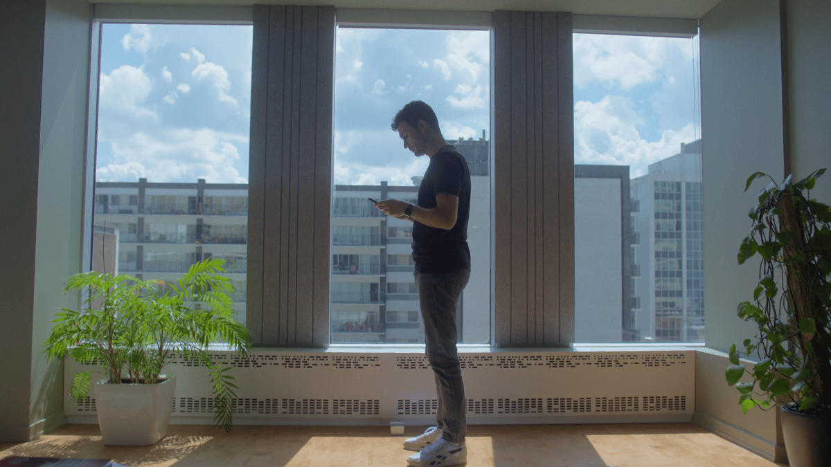 A man stands sideways next to a large glass window in a building, holding a phone with both hands and focused on its screen.