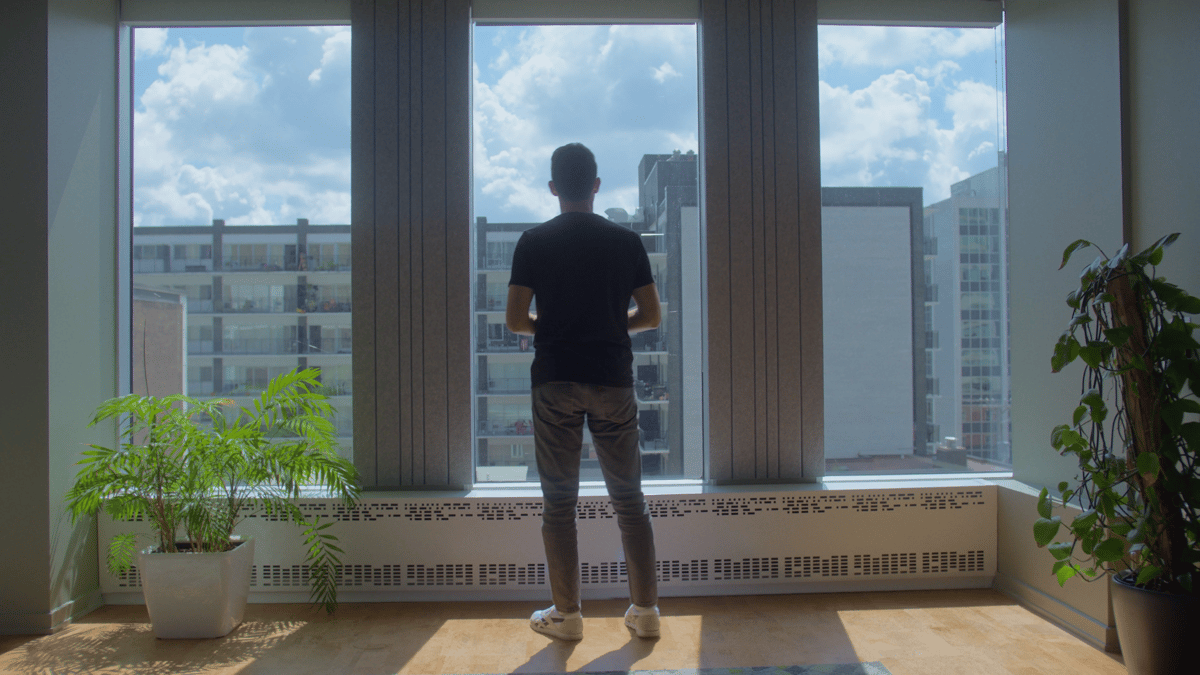 A man stands upright in front of a window, with a view of the building outside.