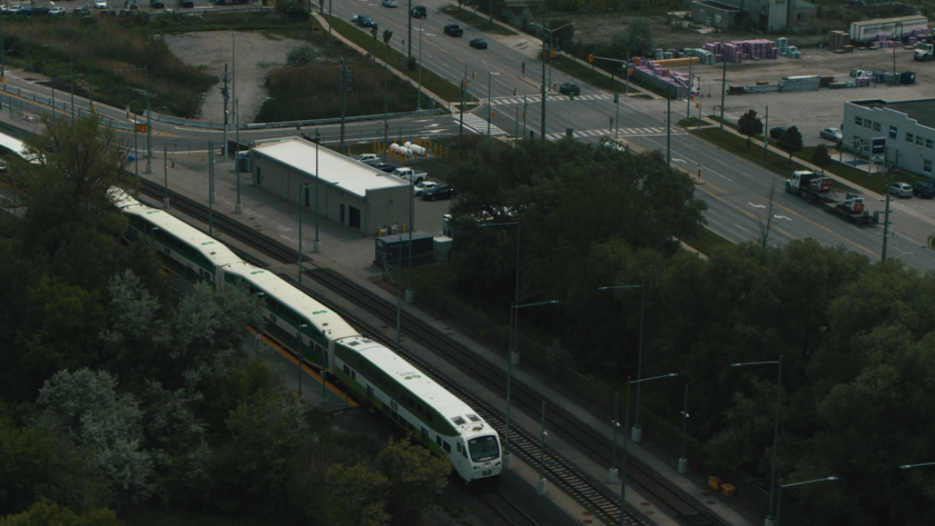 An aerial view of a commuter train pulling into a suburban station surrounded by roads and buildings.