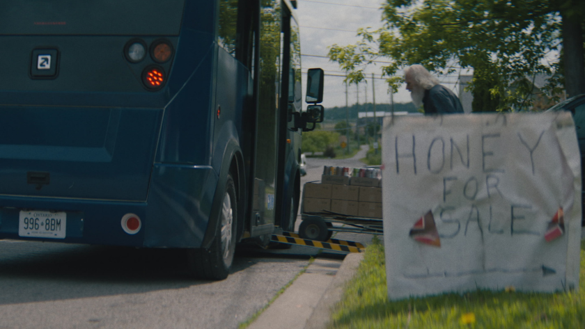 A small transit bus deploys its ramp near a roadside stand with a handwritten "Honey for Sale" sign.