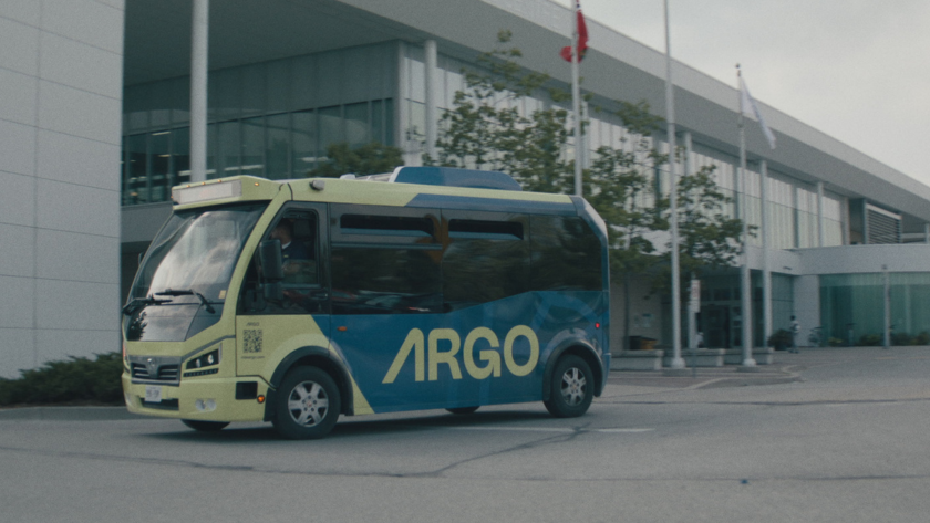 A Ride Argo shuttle bus drives past a modern building with flags at the entrance.