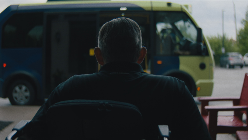 A person in a wheelchair looks toward a green and blue transit bus parked outside.