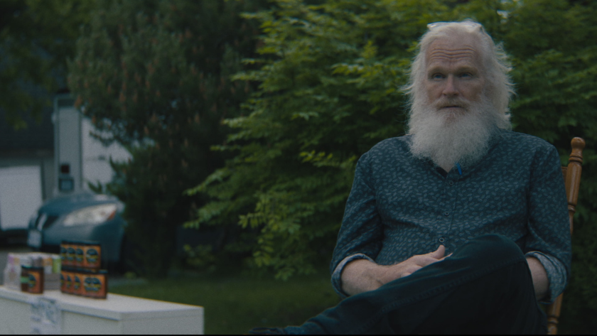 An older man with a white beard sits near a table of honey jars at a roadside stand.