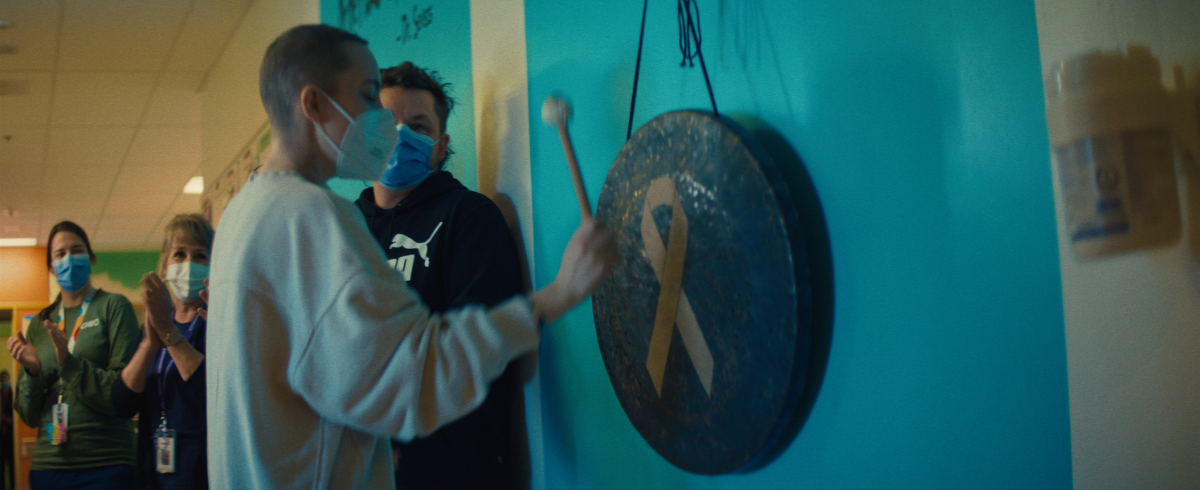 The leukemia patient holding a stick, making an attempt to hit the gong with a yellow ribbon attached to it.