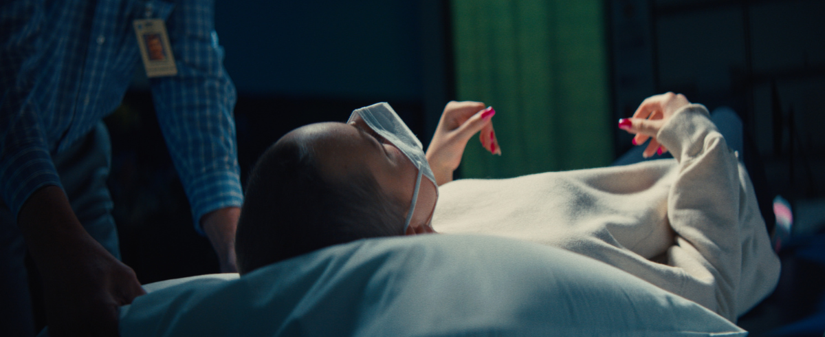 A leukemia patient lying in bed, wearing a white lab gown, while someone is assisting her.