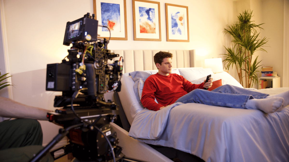 An actor lying down on the bed, looking at a remote while being filmed.
