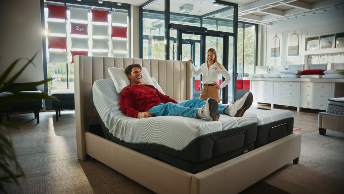 Man lying down in a store while a saleslady stands next to him.