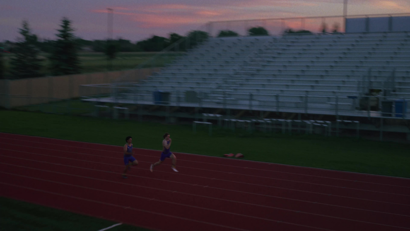Two track athletes sprint side by side on a red running track at sunset.