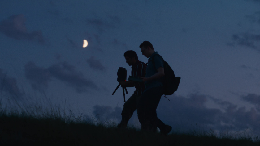 Two students walk under the night sky carrying camera equipment with a crescent moon above.