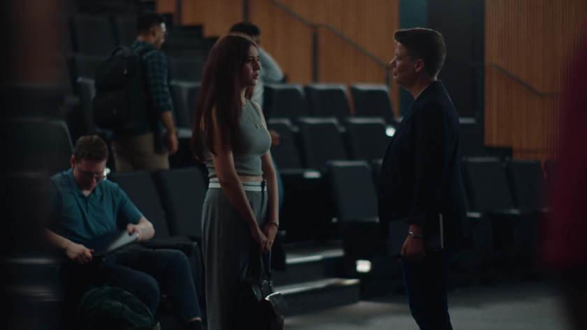 A professor speaks with a student inside a lecture hall as others gather in the background.