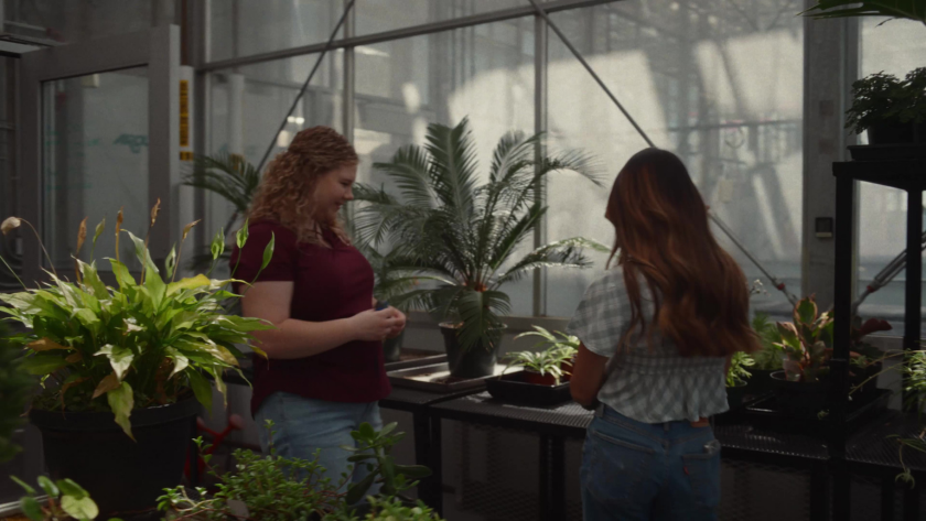 Two students talk inside a greenhouse surrounded by lush green plants.