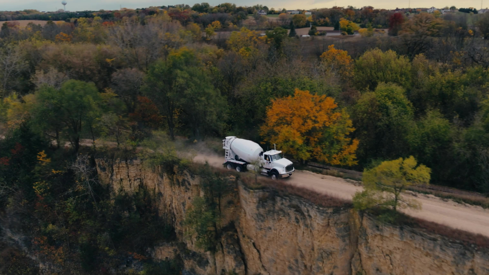 An aerial view captures a concrete mixer truck navigating a winding rural road through autumn foliage, demonstrating commercial vehicle capability in scenic landscape cinematography.