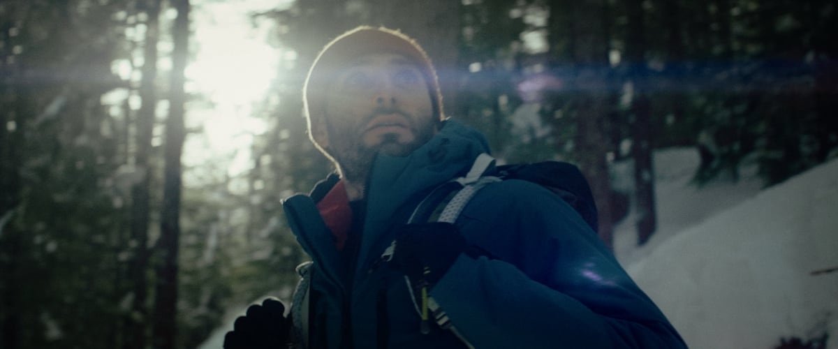  A man looks to his side, clutching the strap of his backpack, surrounded by a forest of tall, magnificent trees, all set against the backdrop of stunning weather.