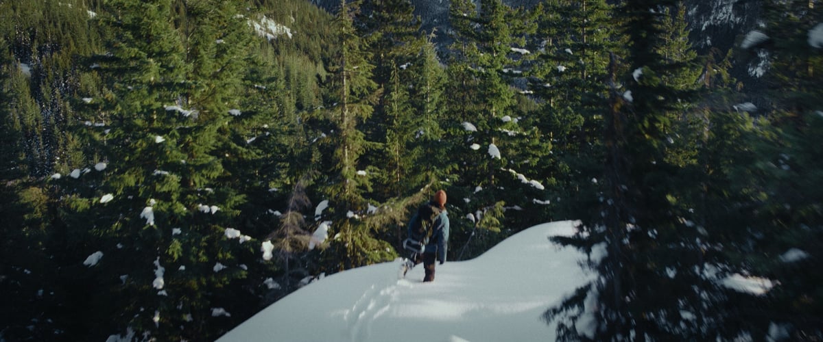 A man strolls through a snowy mountain landscape, surrounded by tall trees, all under the backdrop of beautiful weather.