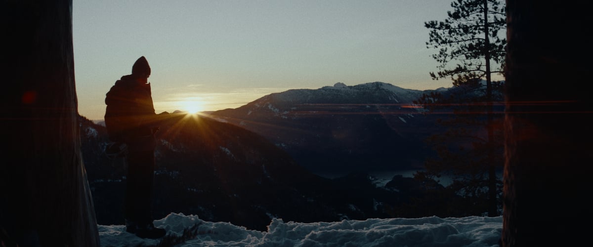  A man stands at the edge of a snowy mountain, bearing witness to a breathtaking sunrise in the majestic mountainous landscape.