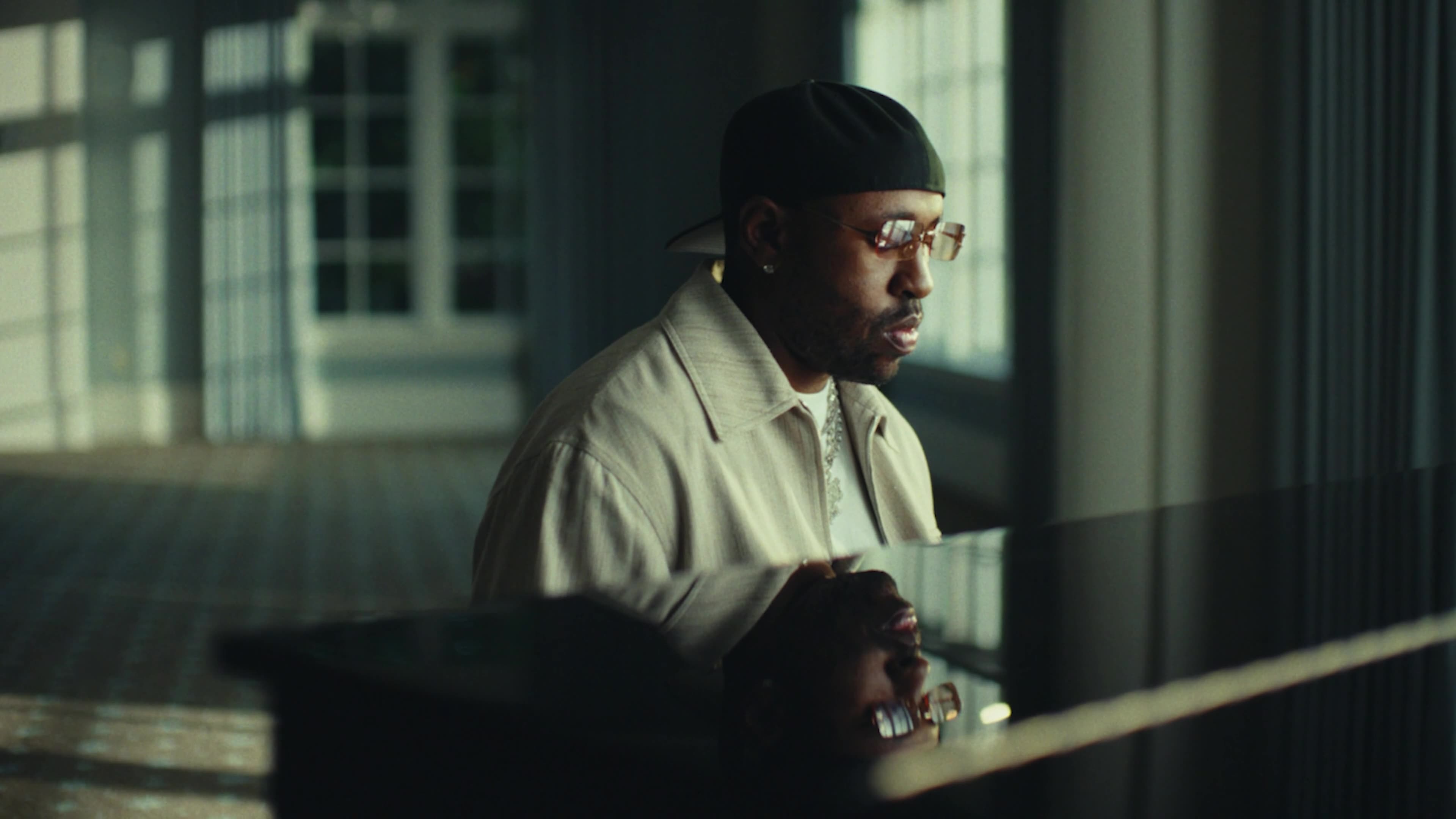 A man in a backwards cap and tinted glasses plays a grand piano in a sunlit room, his face reflected in the polished lid.