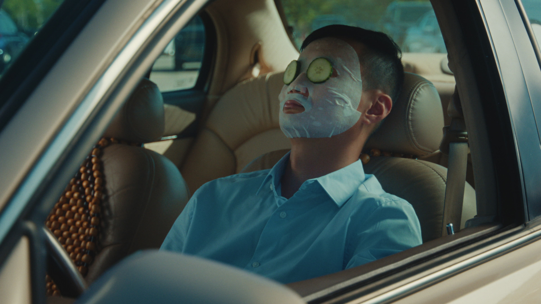 A man relaxes in the drivers seat wearing a sheet mask and cucumber slices, framed through the car window
