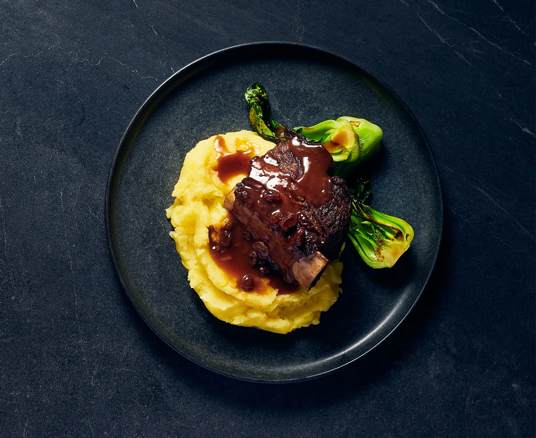 Mashed potatoes with ribeye, gravy, and leeks on a black plate and black background