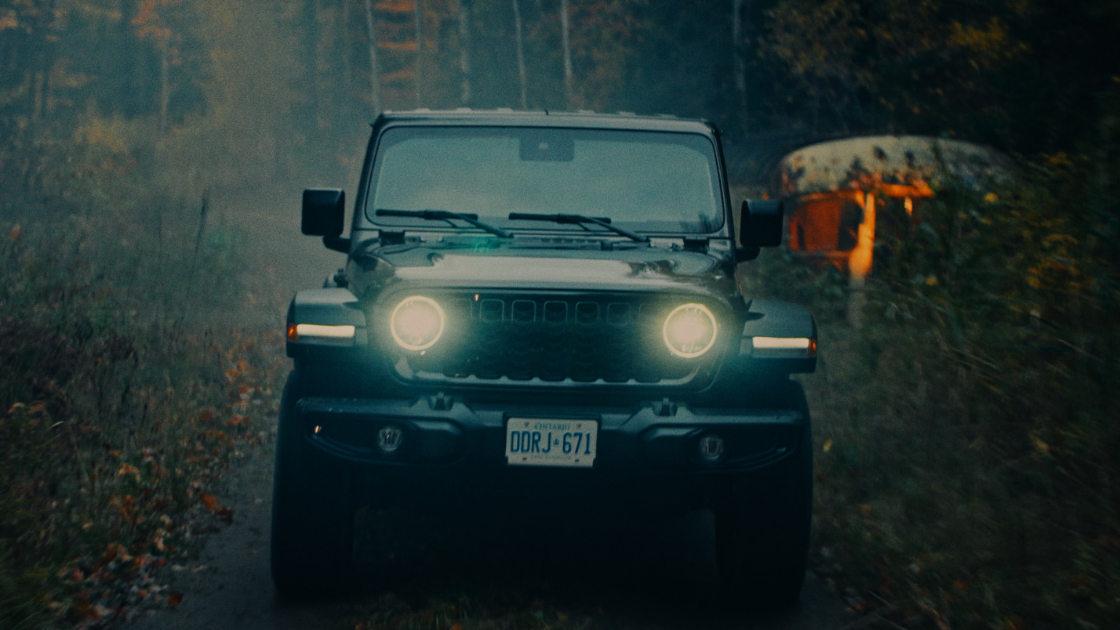 A rugged off-road vehicle drives through a dark, wooded road in rainy conditions, headlights cutting through fog and low light for a cinematic automotive shot.
