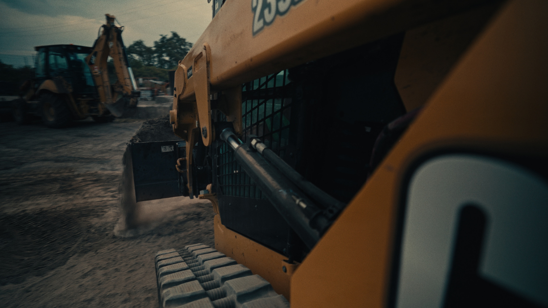 Construction equipment operates on a dirt worksite, with a close-up of hydraulic machinery and moving earth.