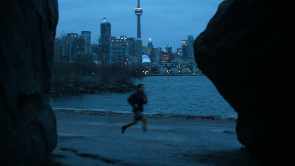 A lone runner jogs along a waterfront path at dusk with the Toronto skyline and CN Tower in the background.
