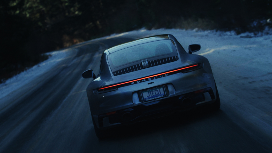 A Porsche sports car drives along a snowy road at dusk, filmed from behind to emphasize speed and motion.