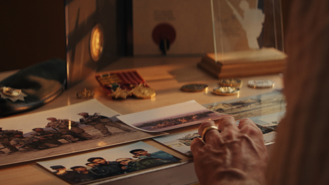 A hand sorts through old printed photographs and military medals on a desk, capturing a reflective moment of memory and service.