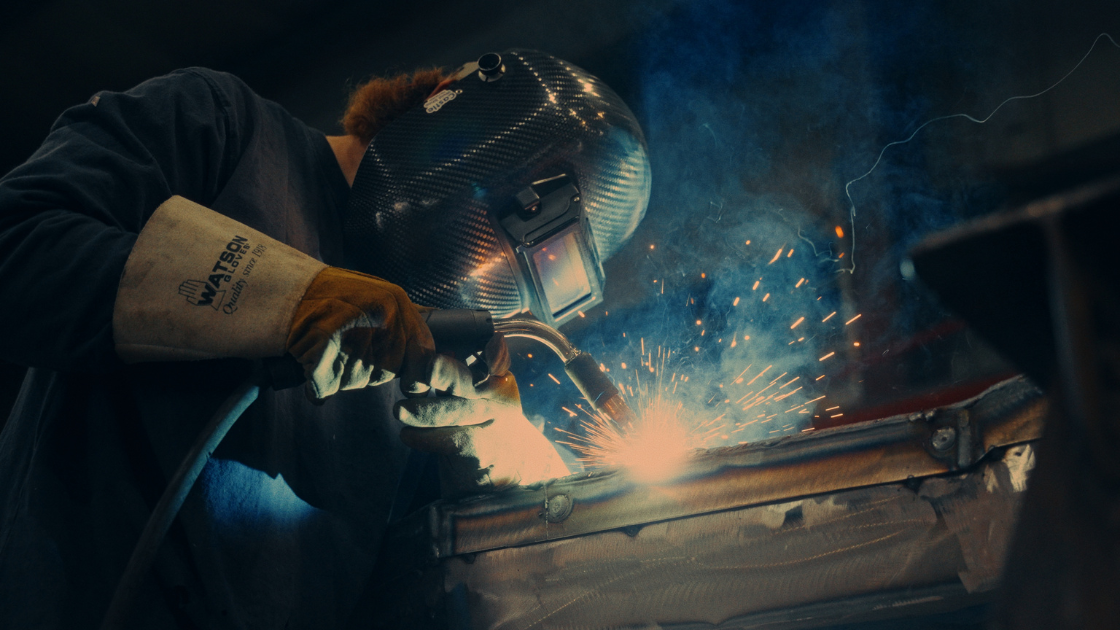 A welder works inside an industrial shop, sparks flying as protective gear and dramatic lighting highlight the craftsmanship.