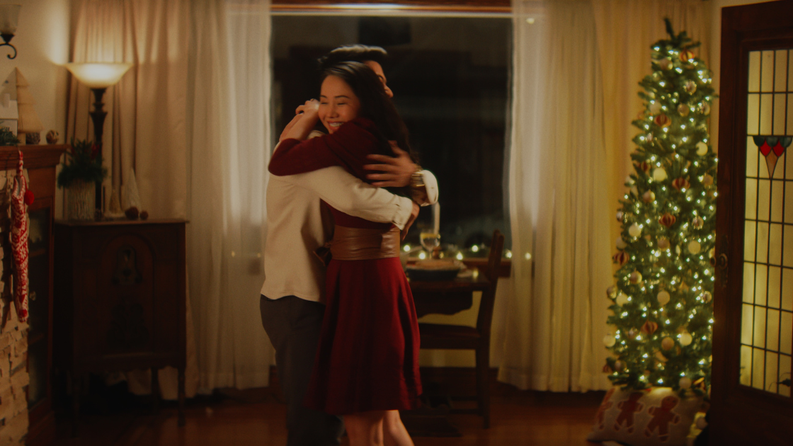 A couple embraces in a warmly lit living room decorated for Christmas, creating an intimate holiday scene.