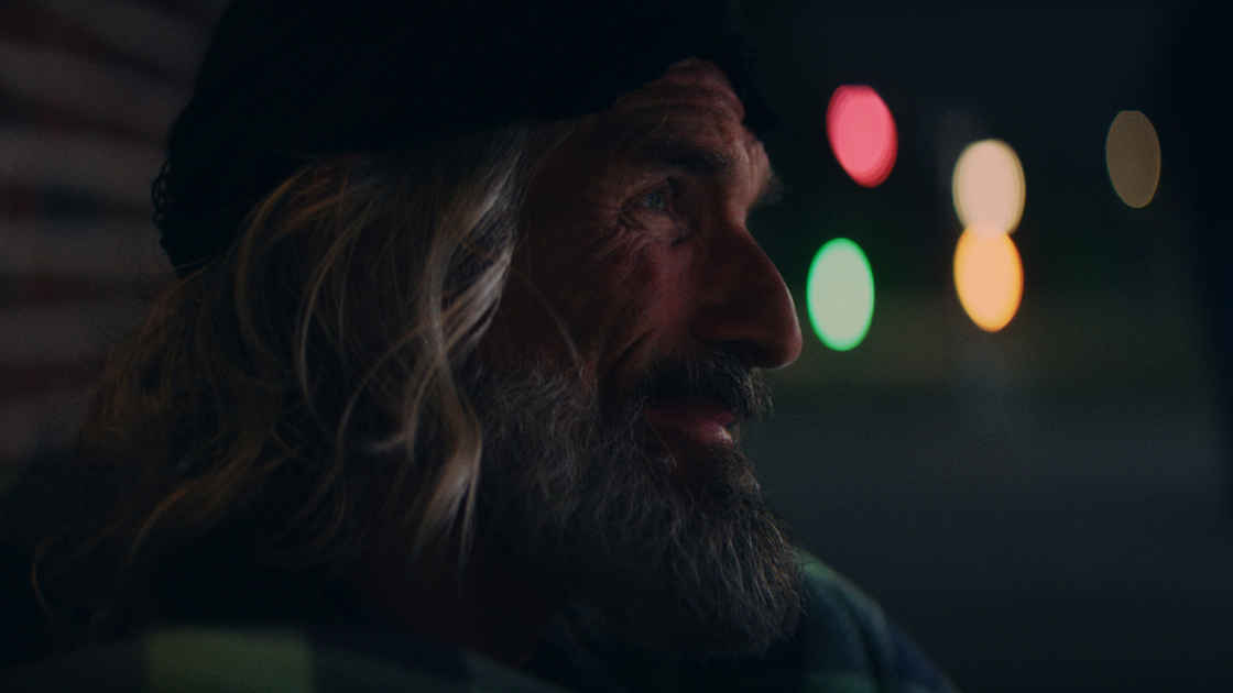 A close-up portrait of an older man with a beard and long hair, lit softly against colorful bokeh lights at night.
