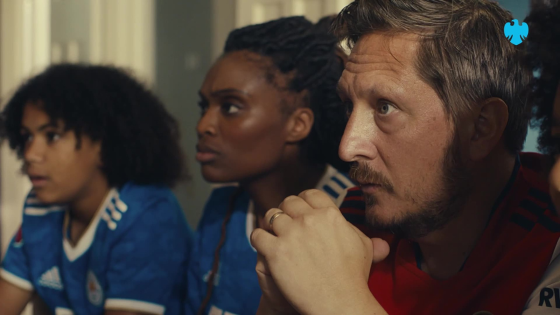 A tense moment unfolds in a cozy living room as a group of football fans—ranging in age and background—watch a crucial match on screen. The close-up framing captures the intense expressions and anticipation. 