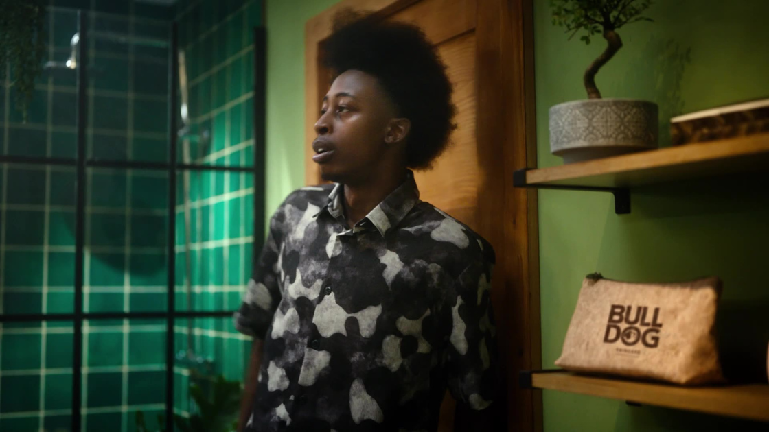 Set in a stylish, plant-filled bathroom with bold green tiles, this image captures a confident young man in a patterned shirt leaning casually against a wall. 