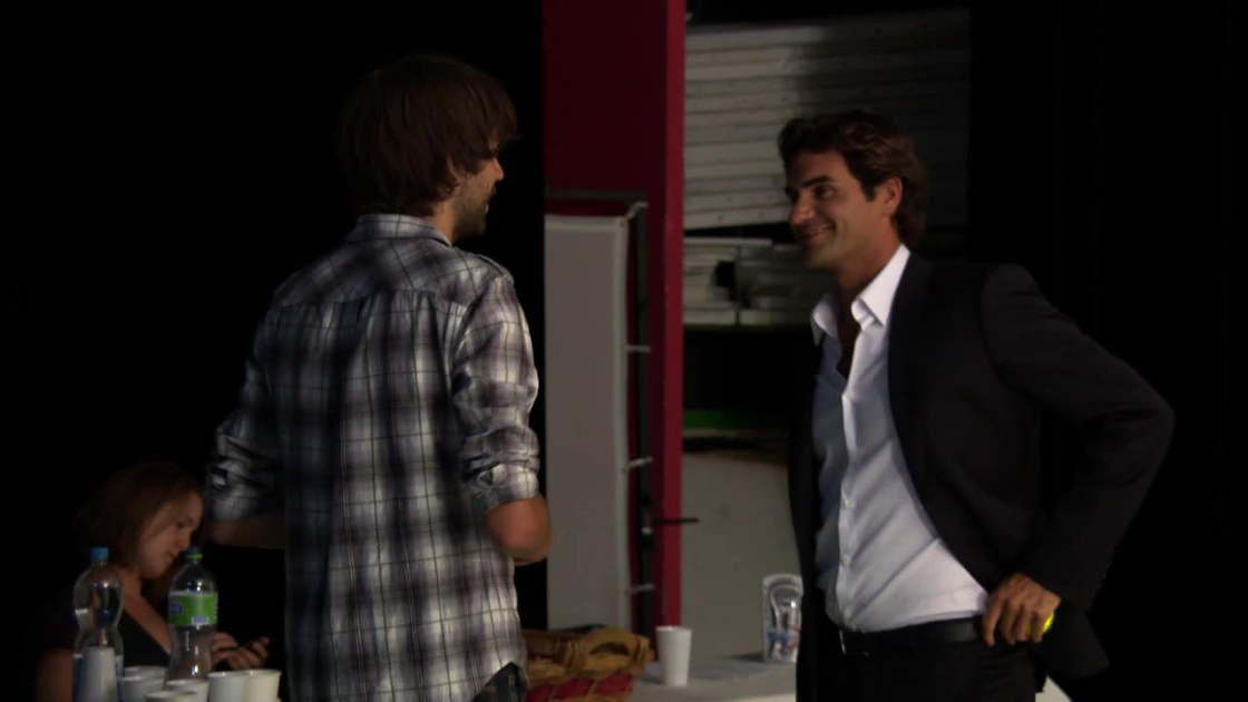 Behind the scenes on set, a casually dressed crew member chats with tennis legend Roger Federer, who stands relaxed in a sharp black suit and white shirt. 