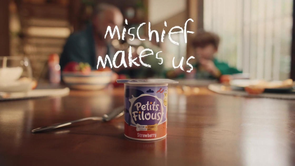 A brightly lit breakfast scene frames a single yogurt pot of Petits Filous in sharp focus on a wooden table. Behind it, a joyful, slightly chaotic family breakfast is hinted at in soft blur.