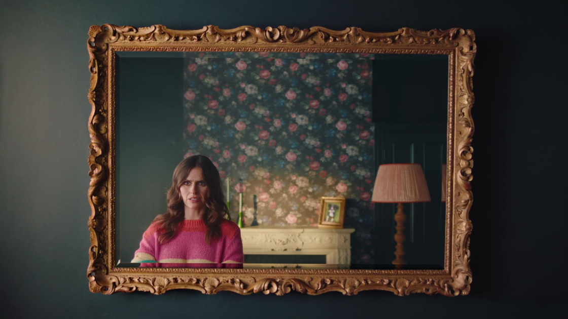 An elegant woman appears framed in a golden antique mirror, situated in a richly decorated living room with floral wallpaper. 
