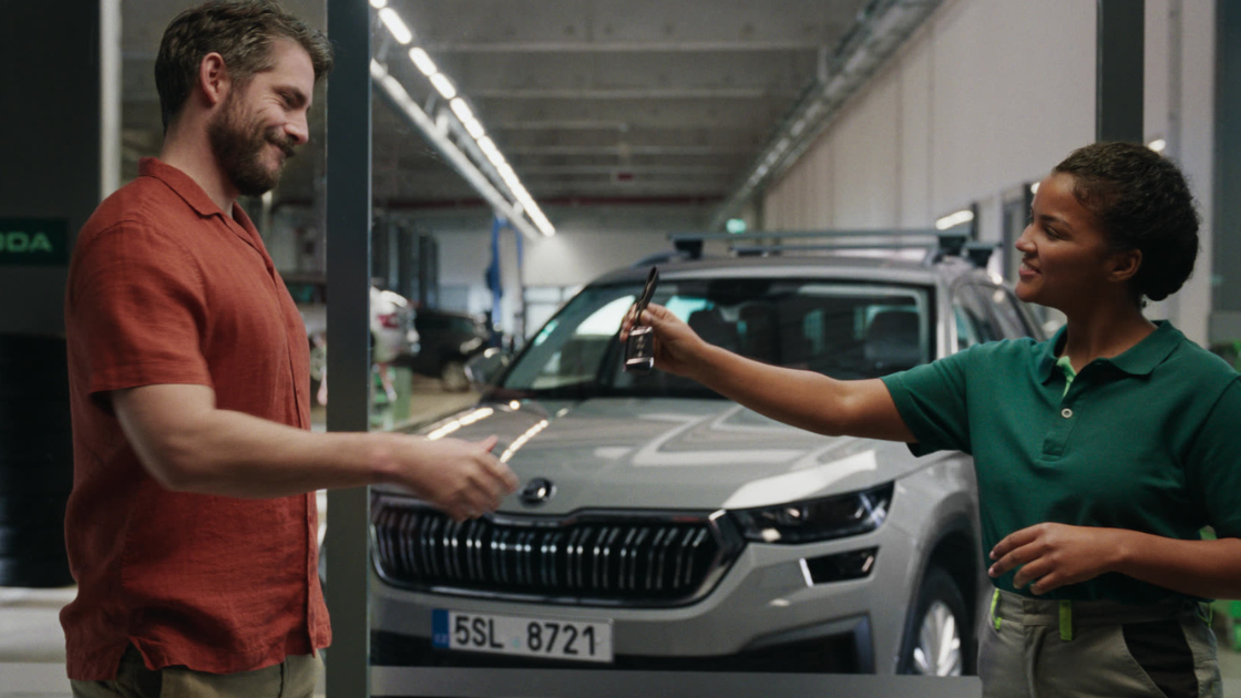 In a bright, clean car dealership, a young woman in a mechanic’s uniform hands over car keys to a smiling customer. 