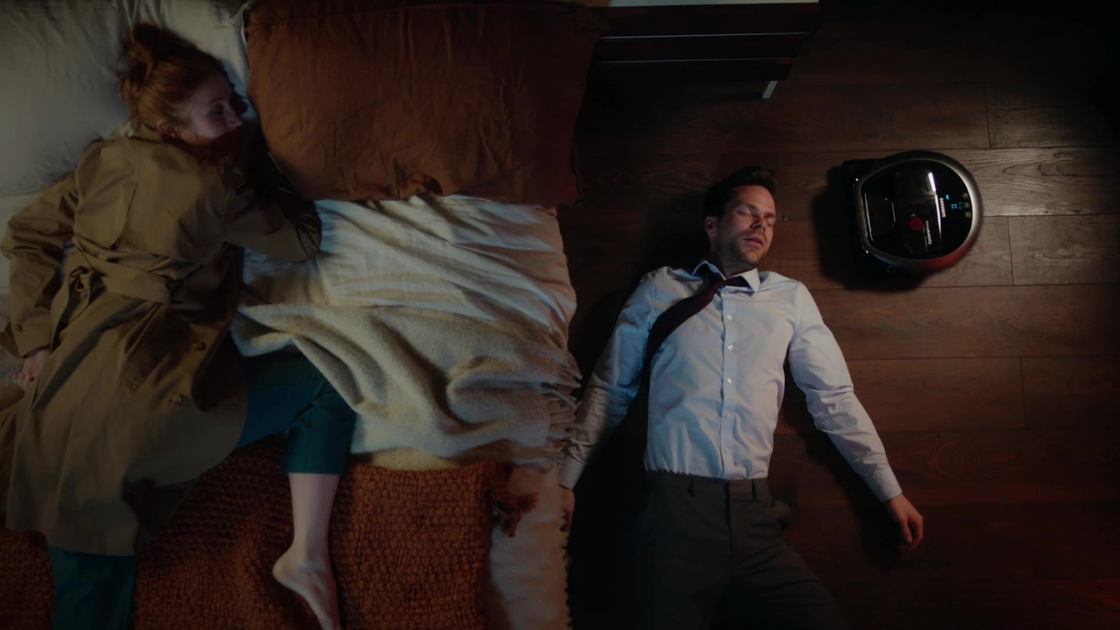 Two professionals are seen collapsed in exhaustion beside a stylish bed, still in work attire, while a sleek robotic vacuum cleaner roams the floor. 