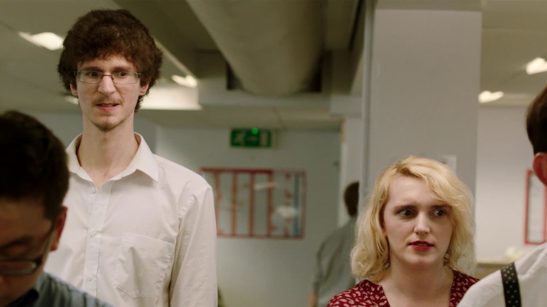 In a brightly lit office environment, two employees stand out amid the bustle—one notably tall man in a white shirt and a blonde woman in red patterned attire. 