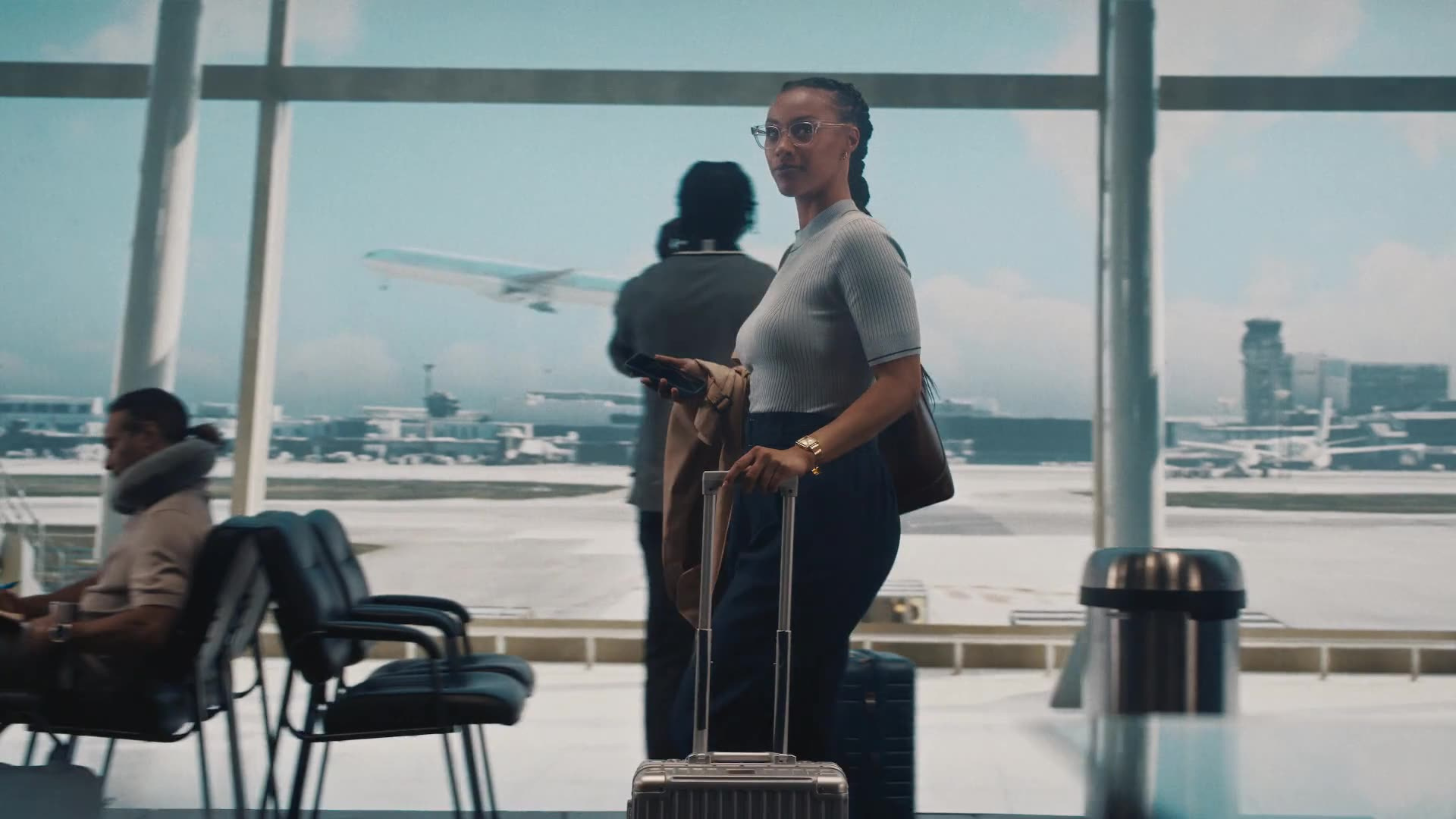 A woman with braids and glasses stands with a rolling suitcase in an airport terminal as a plane takes off outside.