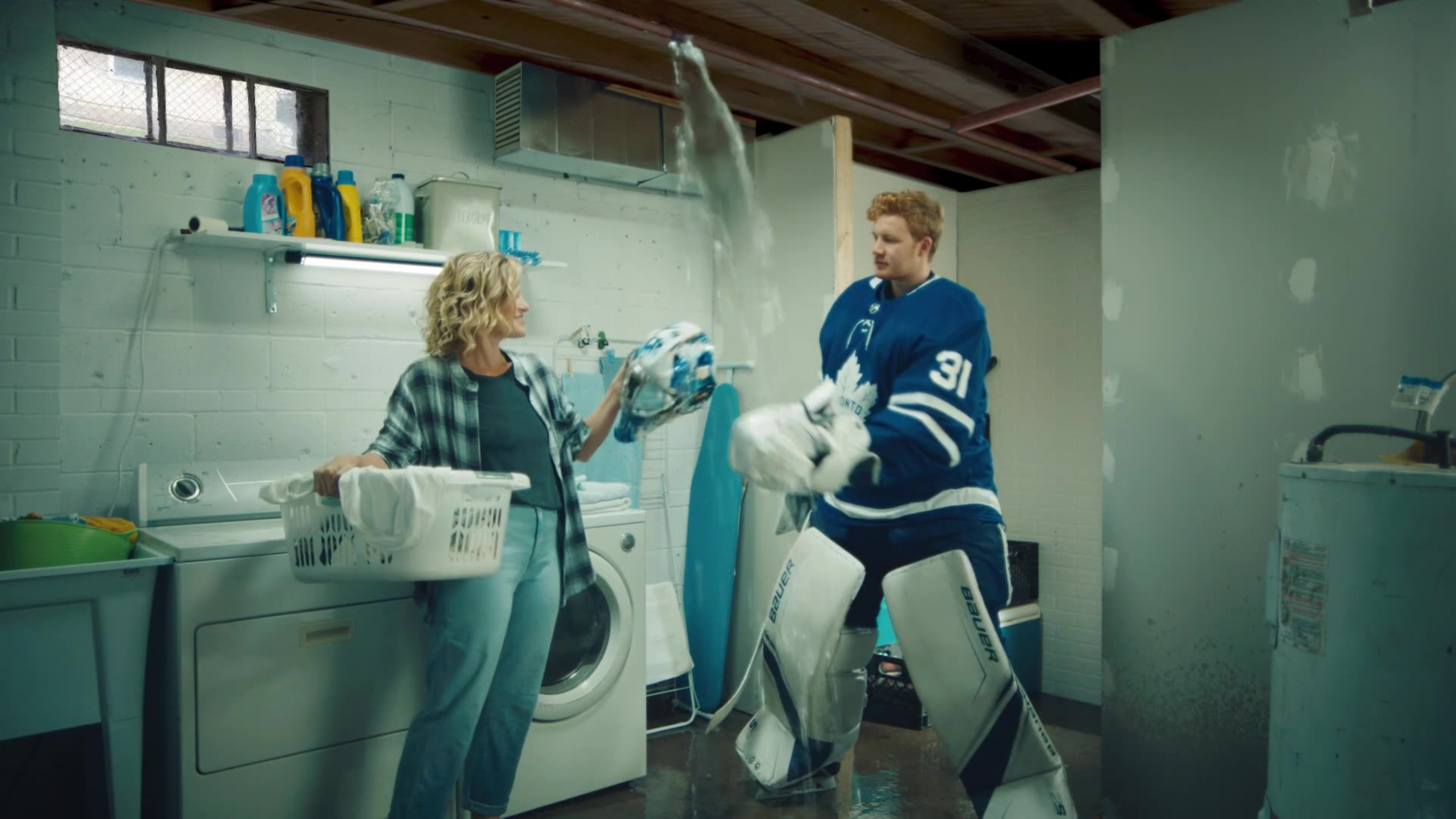 A young man in a Toronto Maple Leafs goalie uniform tosses laundry with a woman in a basement laundry room.