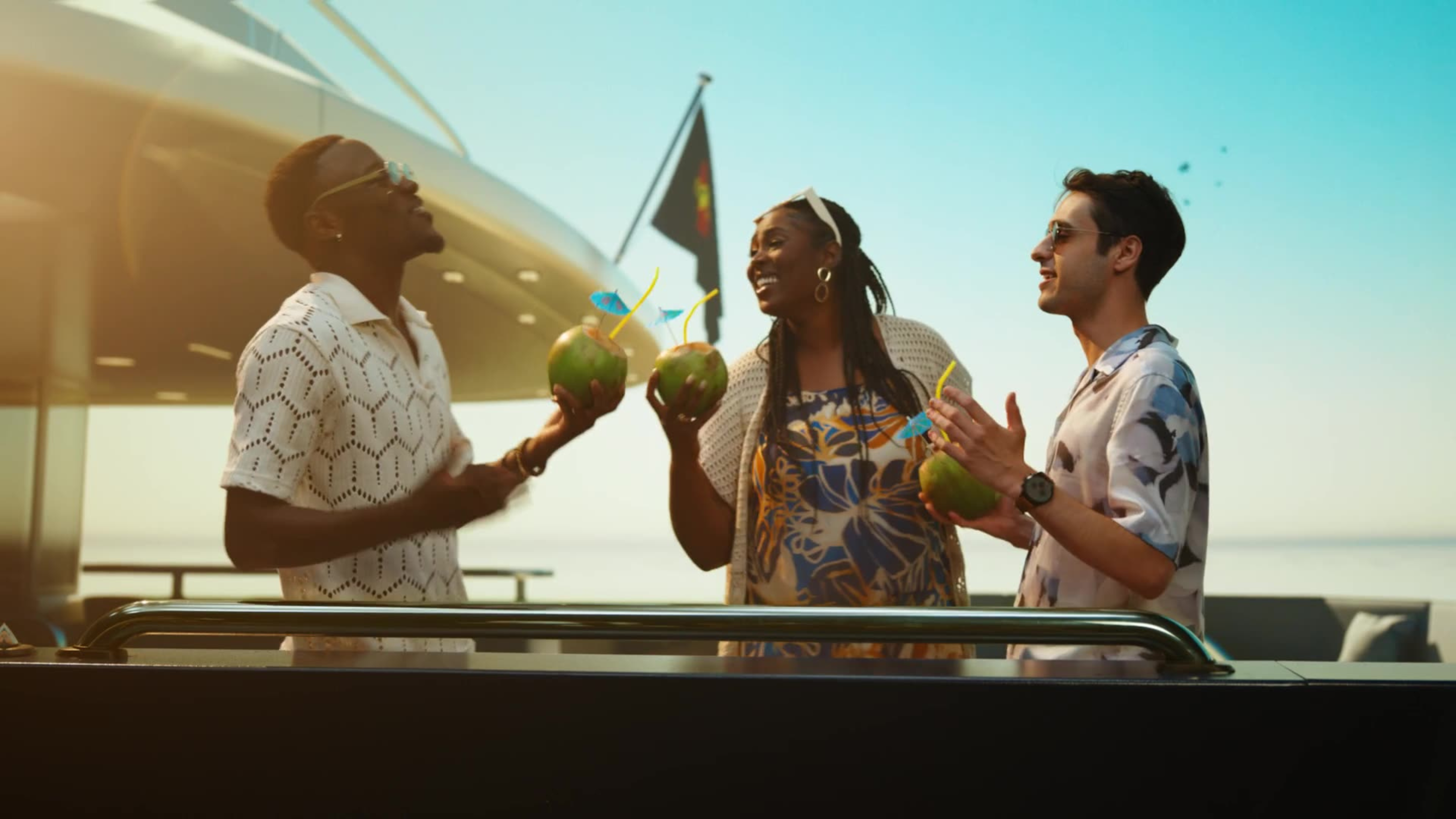 Three friends laugh and clink coconut drinks together on the deck of a luxury yacht.