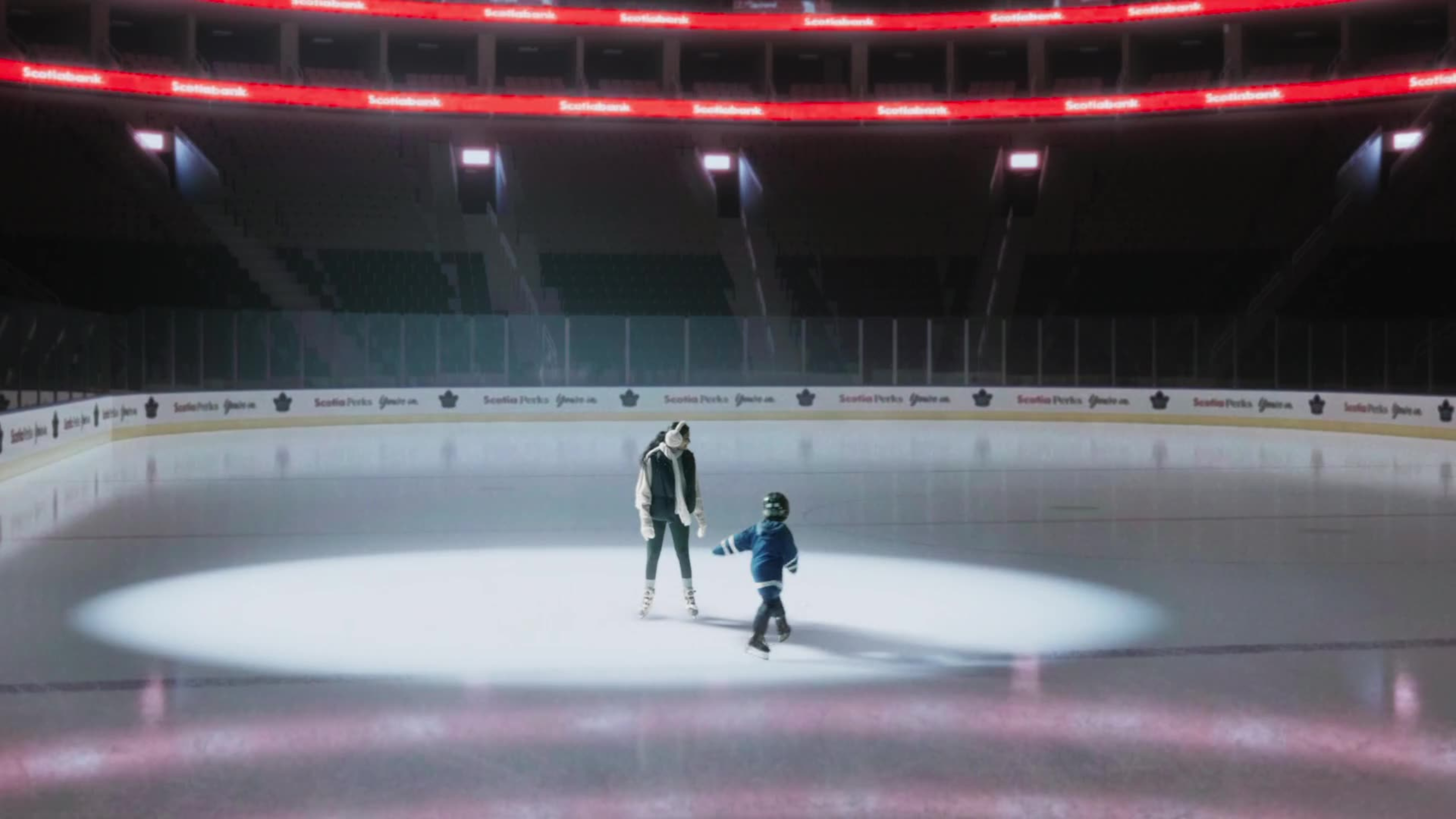 A parent and small child in a Leafs jersey skate together under a spotlight on the empty Scotiabank Arena ice.