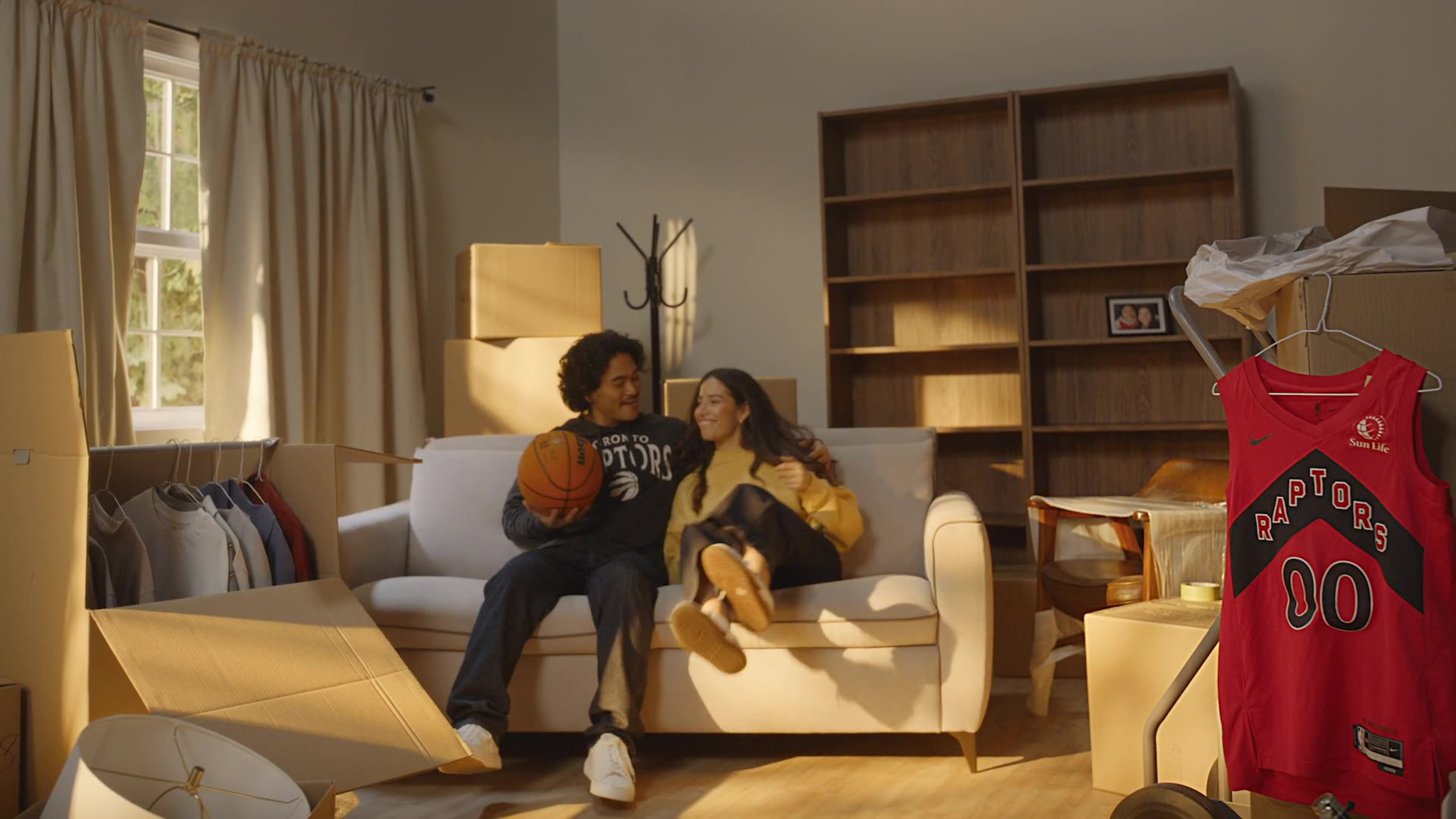 A couple in Raptors gear laugh together on a sofa surrounded by moving boxes, with a red Raptors jersey hanging nearby.