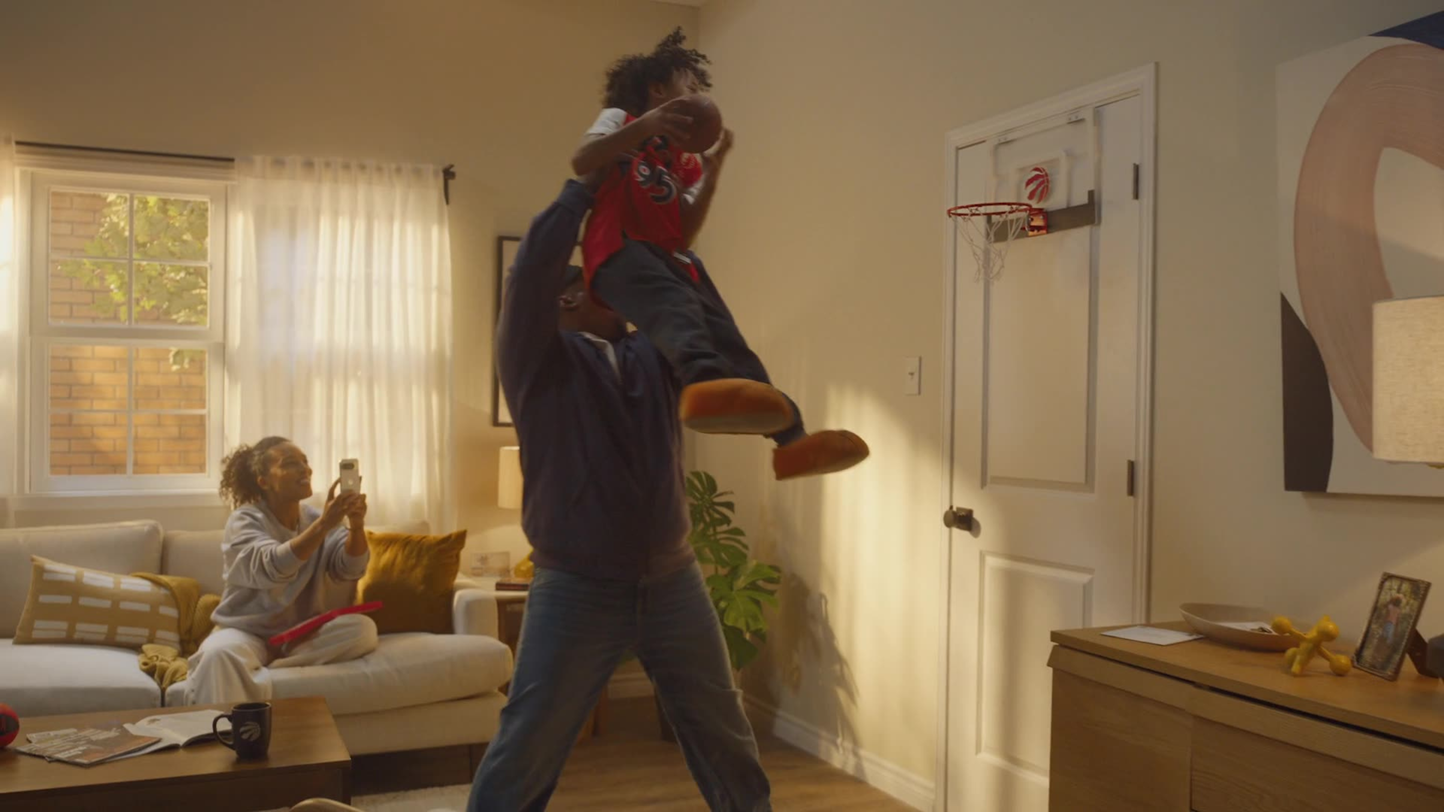 A father lifts a small child in a Raptors jersey toward a mini basketball hoop on the door while a woman films from the couch.